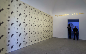 Vista de una sala de la exposición Segunda vez de Dora García. Junto al muro empapelado con el lacaniano papel pintado se sitúa una obra de la serie Golden Sentences. Al fondo, la película "La lección respiratoria".
