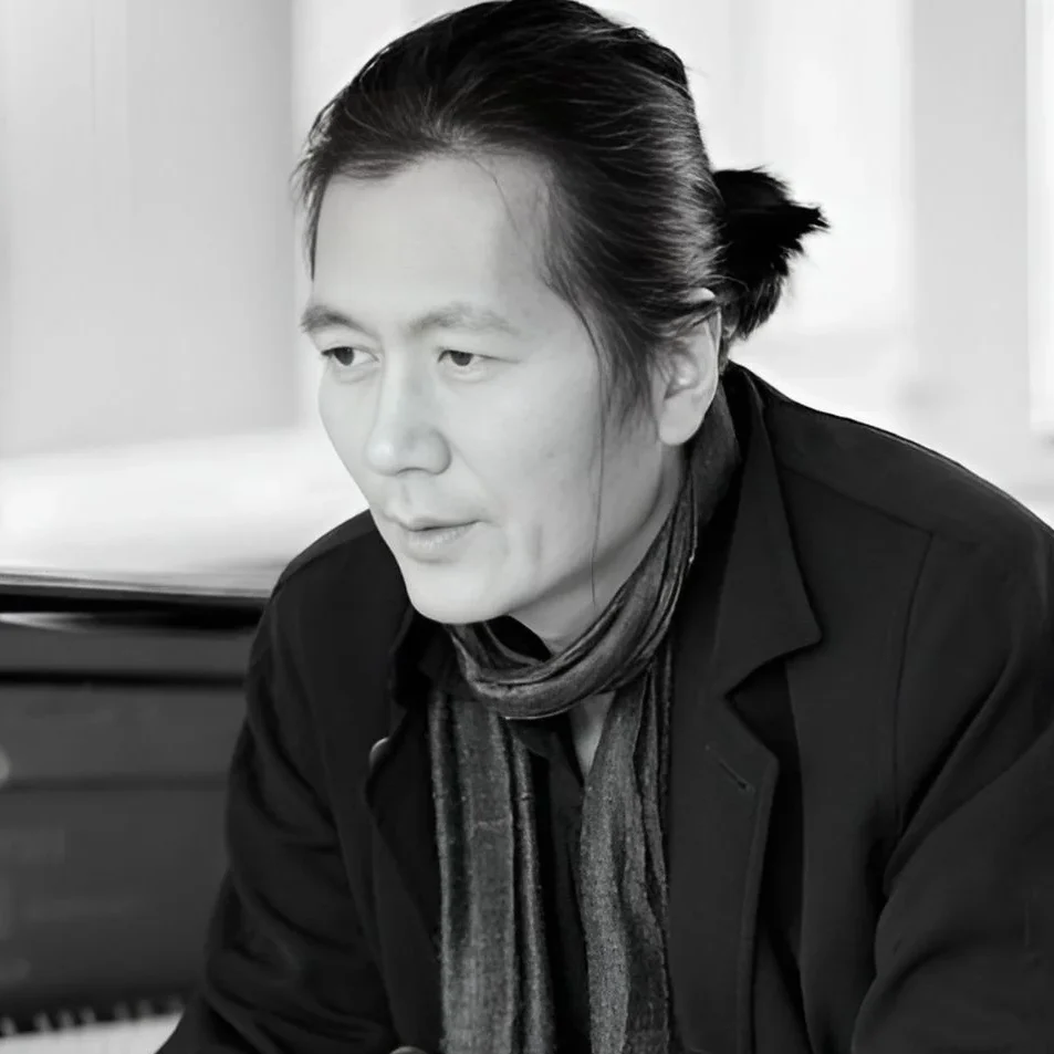 Fotografía de Byung-Chul Han. Muestra a un hombre de rasgos asiáticos, con el pelo negro y liso recogido en una coleta corta baja. Está sentado delante de un piano y mira hacia el suelo lejano a la izquierda de la cámara, con gesto pensativo, como haciendo una pausa mientras habla.