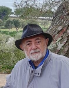 Fotografía de Carlos García Gual. Muestra a un hombre mayor de rasgos caucásicos que mira a cámara con expresión afable sentado en un árbol, con un campo de árboles bajos de fondo. Tiene una barba blanca de longitud media que le cubre la mandíbula y la boca, y lleva un cortavientos blanco y un sombrero fedora azul oscuro.