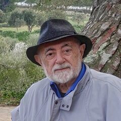 Fotografía de Carlos García Gual. Muestra a un hombre mayor de rasgos caucásicos que mira a cámara con expresión afable sentado en un árbol, con un campo de árboles bajos de fondo. Tiene una barba blanca de longitud media que le cubre la mandíbula y la boca, y lleva un cortavientos blanco y un sombrero fedora azul oscuro. 